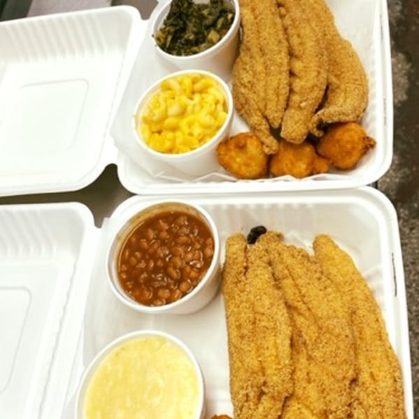 Two takeout containers of David's Catfish House fried catfish, hushpuppies, mac and cheese, greens, baked beans, and grits.