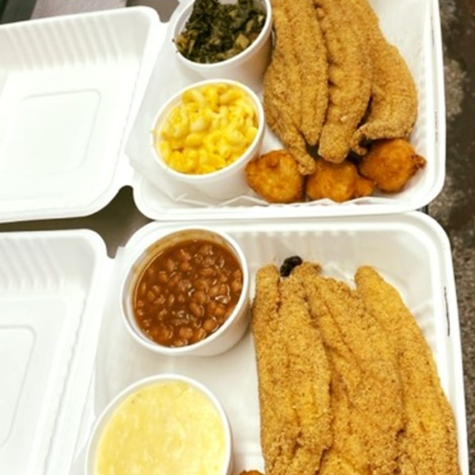 Two takeout containers of David's Catfish House fried catfish, hushpuppies, mac and cheese, greens, baked beans, and grits.